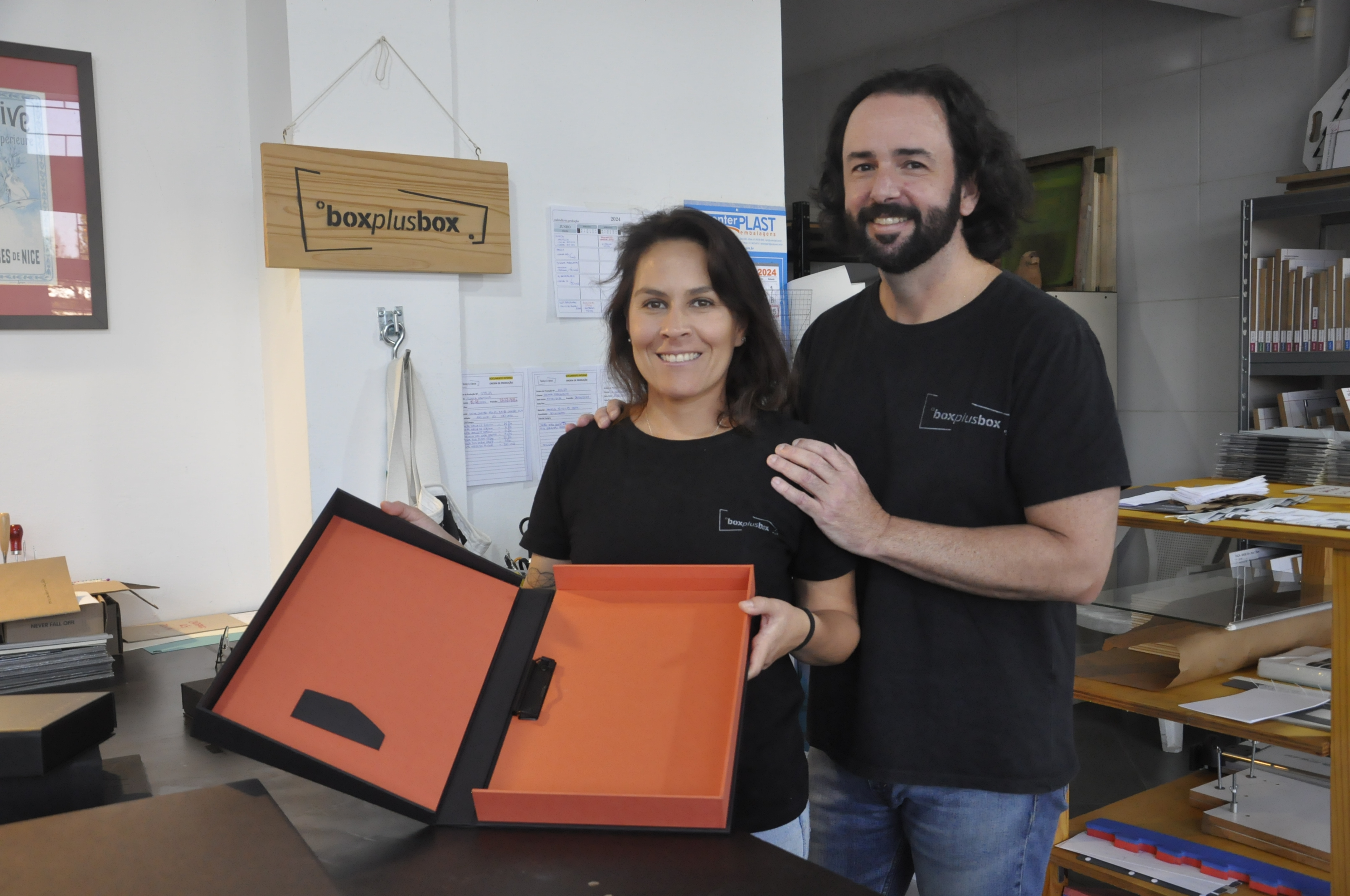 Patrícia Motta e Daniel Cortezia são jovens, brancos, de cabelos escuros, ele é mais alto e usa barba e cabelos compridos. Eles vestem camisetas pretas e mostram produtos da cartonagem que montaram com recursos da Fomento Paraná