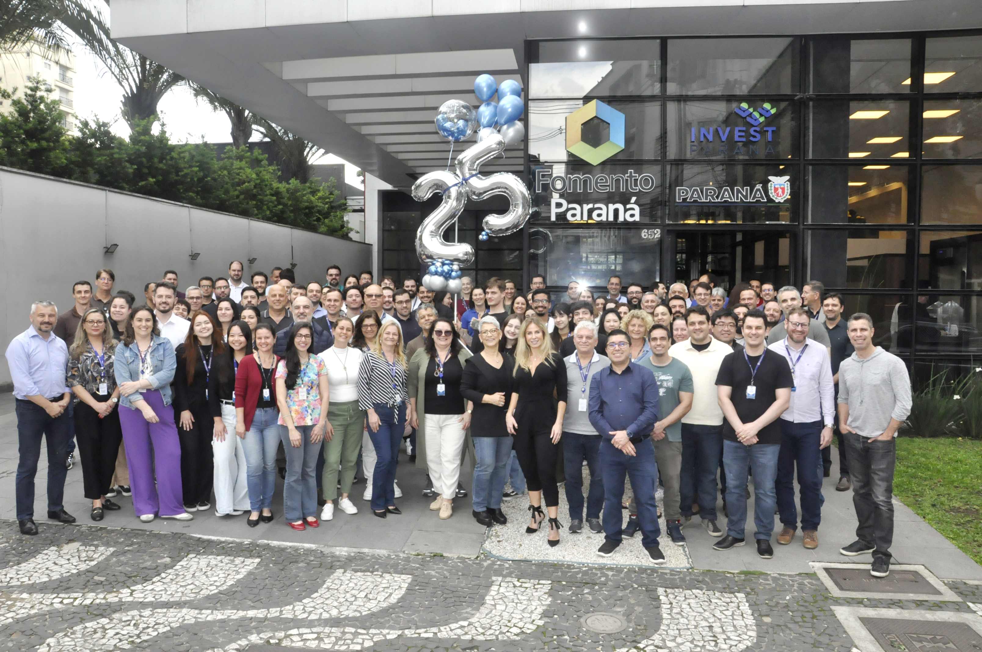 A imagem mostra grupo de mais de 100 colaboradores da Fomento Paraná posando para foto em frente à empresa